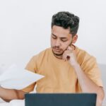 Un jeune homme d'affaires réfléchi examine des documents tout en travaillant sur un ordinateur portable à l'intérieur.