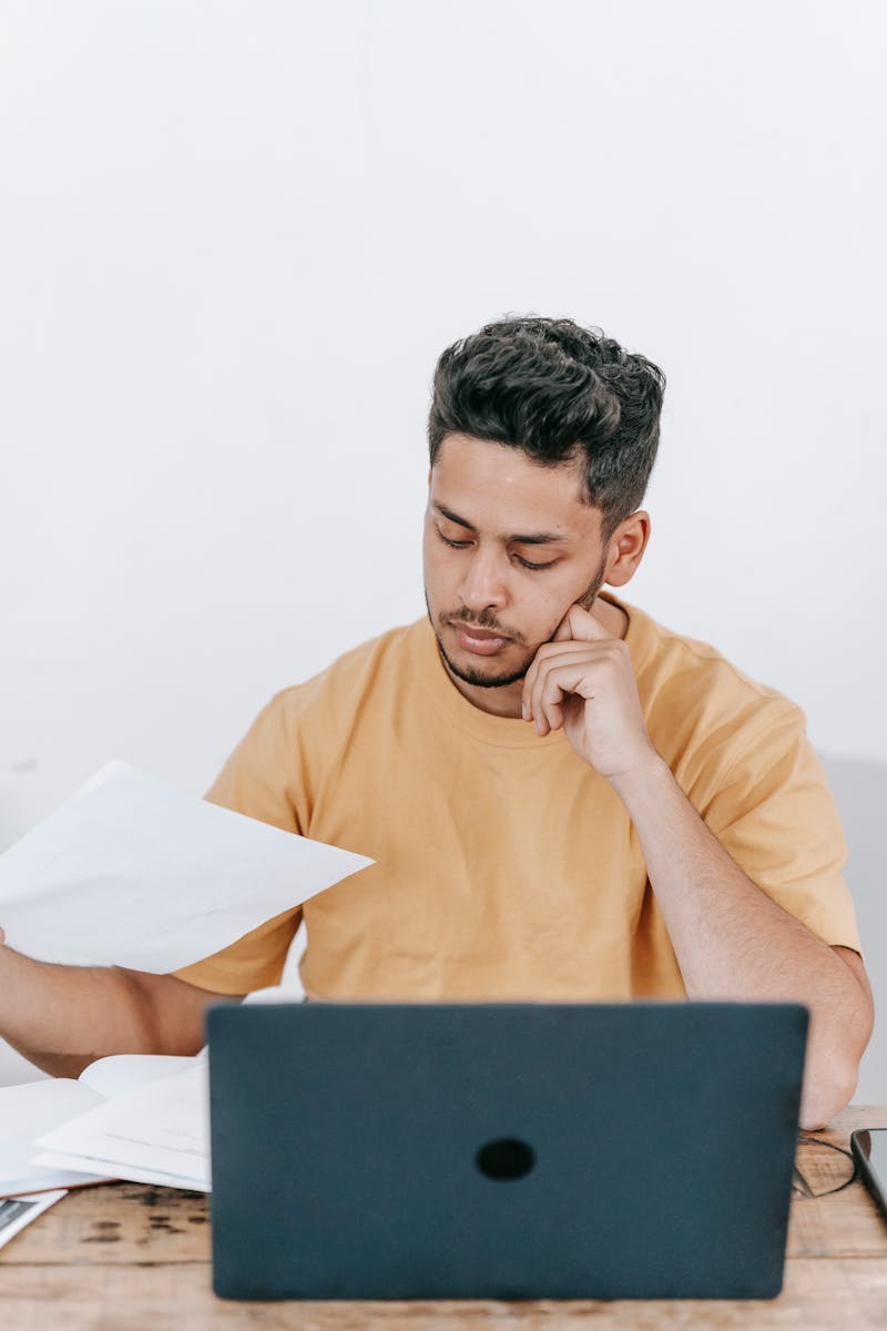 Un jeune homme d'affaires réfléchi examine des documents tout en travaillant sur un ordinateur portable à l'intérieur.
