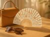 Un sac en paille, des lunettes de soleil et un éventail de bois à motifs floraux posés sur une table, illustrant L’Éventail de Poche idéal pour l’été.