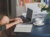 Une femme travaille sur ordinateur, note dans cahier, boit café près de fleurs