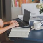 Une femme travaille sur ordinateur, note dans cahier, boit café près de fleurs