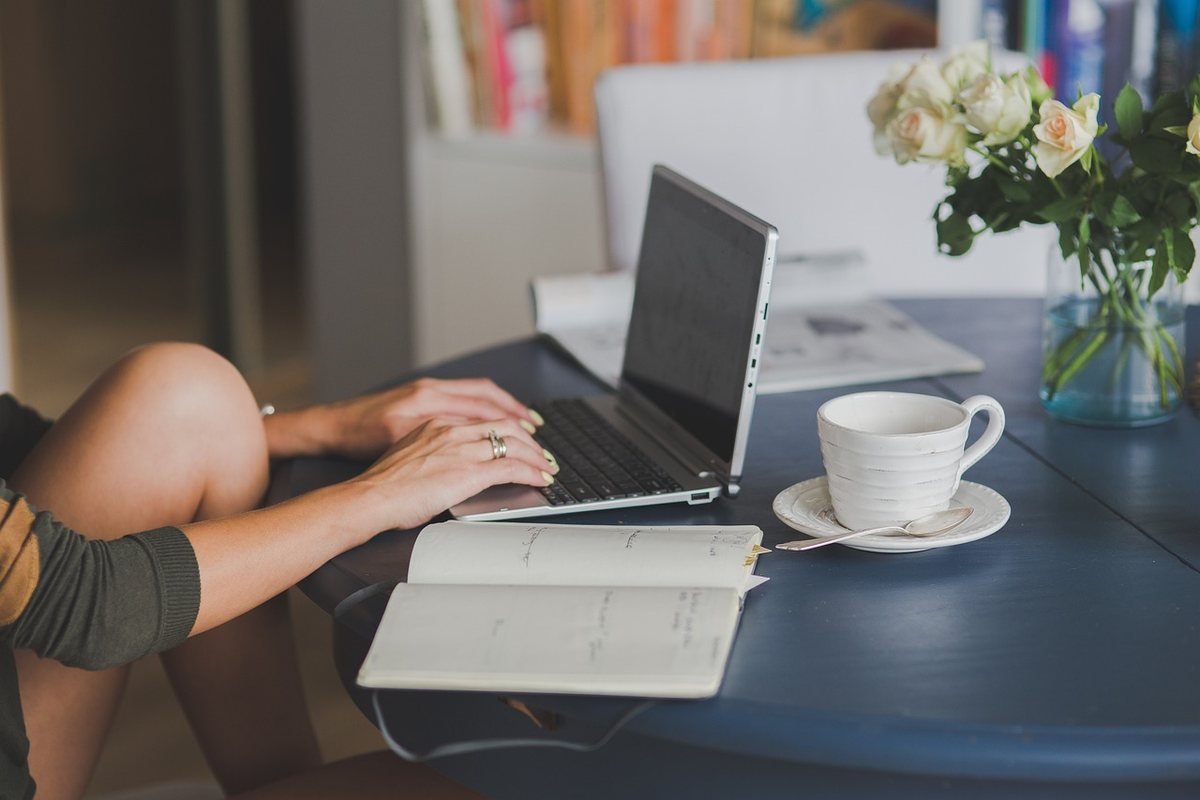 Une femme travaille sur ordinateur, note dans cahier, boit café près de fleurs